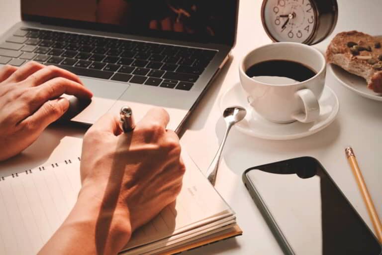 A man's hand writing in a notebook, near a laptop computer and coffee