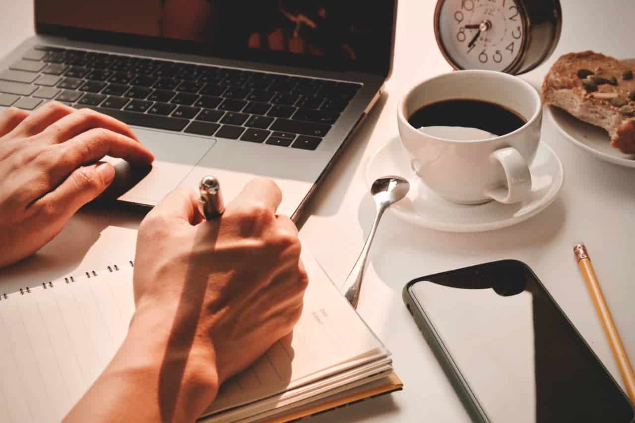 A man's hand writing in a notebook, near a laptop computer and coffee